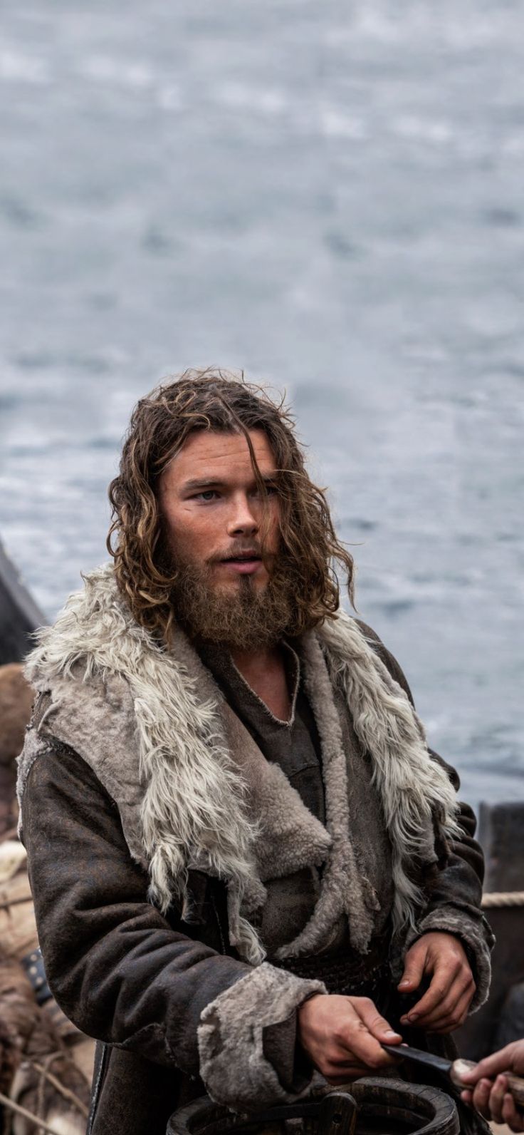 a man with long hair and beard wearing a fur lined coat on the deck of a boat