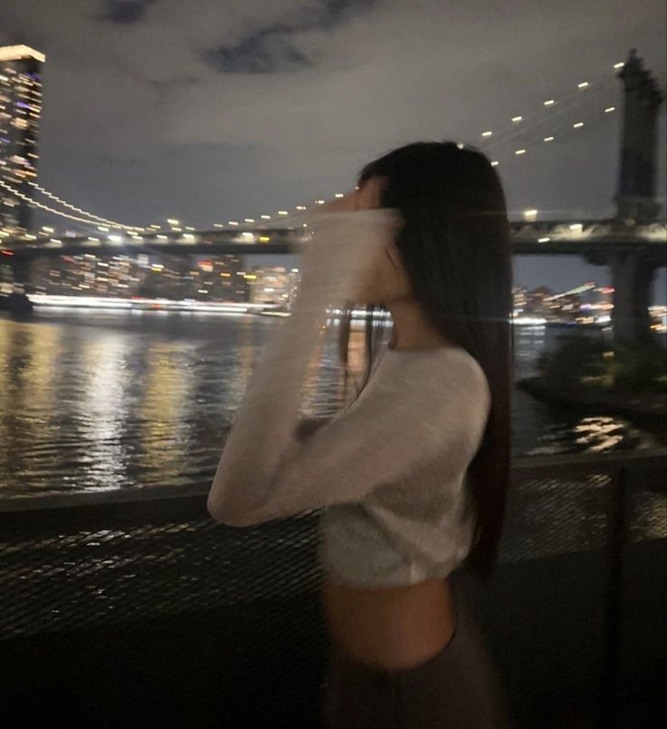 a woman standing in front of a river at night with the city lights behind her