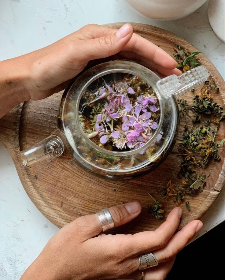 two hands holding a glass container filled with flowers on top of a wooden tray next to a cup