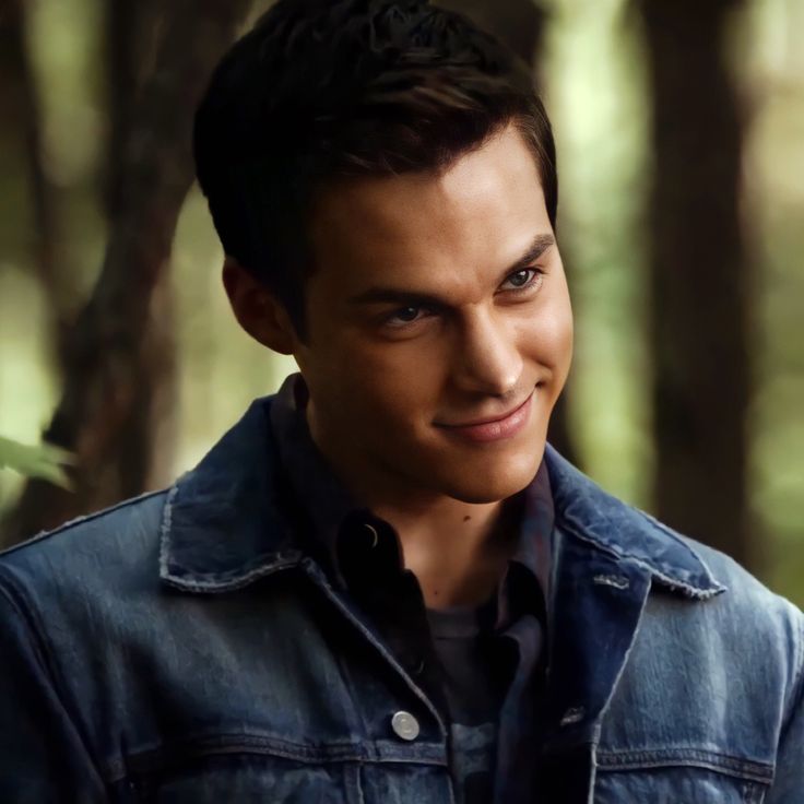 a young man wearing a jean jacket standing in the woods looking at the camera with a smile on his face