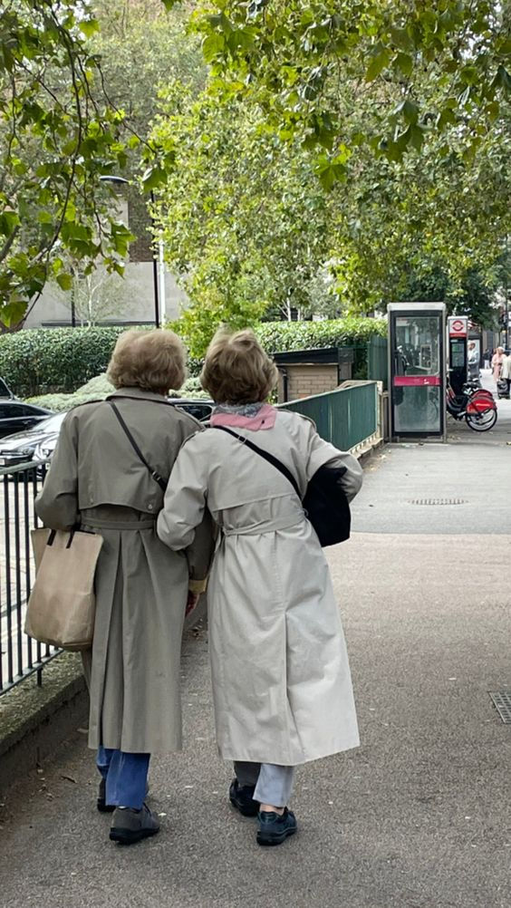 two people are walking down the sidewalk near a fence and trees, one is wearing a gray coat