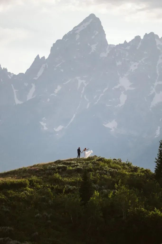 two people standing on top of a lush green hillside with snow covered mountains in the background