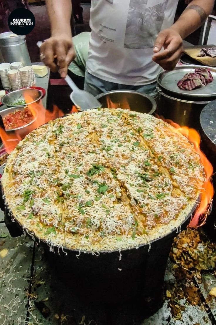 a man standing over a large pizza on top of a fire pit in front of other food