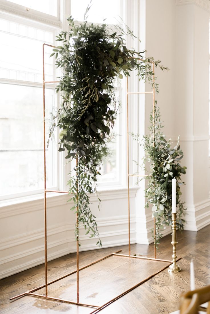 two gold stands with greenery on them in front of a white wall and window