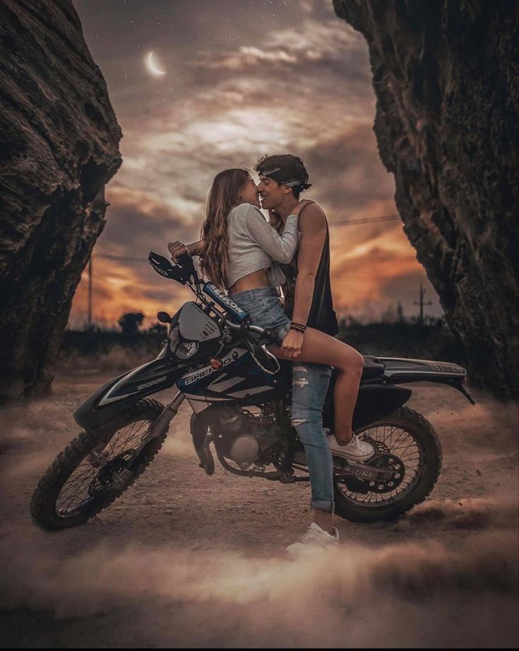 a man and woman sitting on a motorcycle in front of some rocks at night with the sun setting behind them