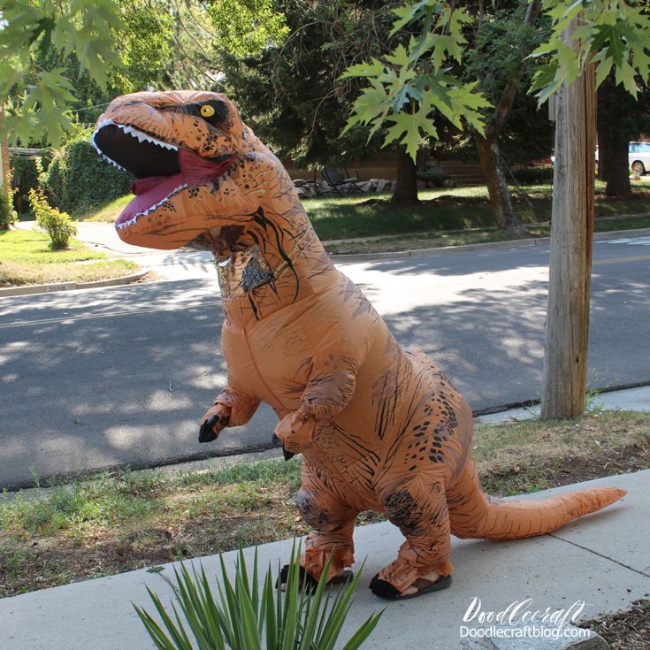 an inflatable t - rex is standing on the sidewalk next to a plant