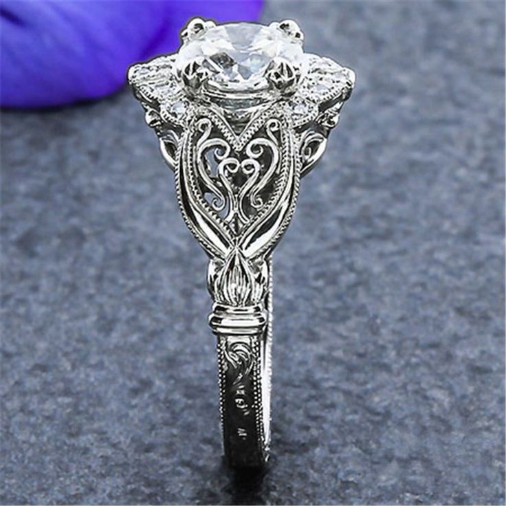 an antique style diamond ring with filigrees and scrolls on the sides, sitting next to a purple flower