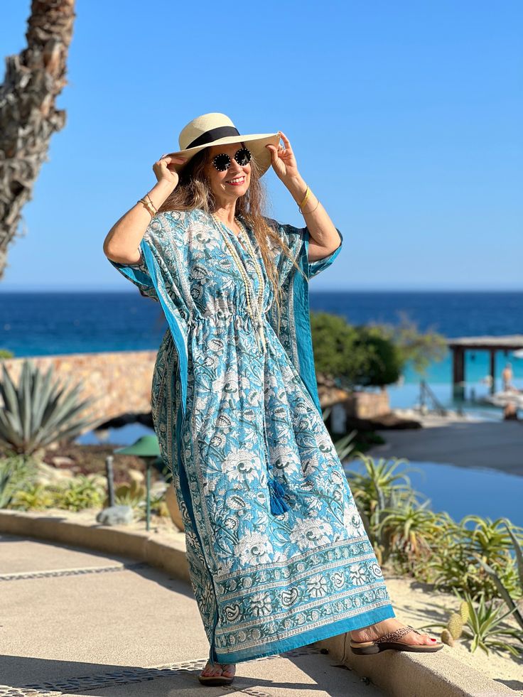 Our Aegean Blue Floral Maxi is an all over floral kaftan dress that is the perfect piece to dress up and wear out, wear to the beach as a cover-up, or just lounge about in it at home! This beautiful dress is named after none other than the aegean blue sea, which boasts the most gorgeous blue/teal colouring! Subtle variations in colour and pattern are to be celebrated as they are evidence of the hand made block printing and dyeing process that makes this dress unique. This dress is the epitome of classic comfort and is perfect to use spring, summer, and into early fall. It is also available in midi and short lengths. One size with drawstring waist Length is 54" 100% cotton voile Made in India Machine wash delicate cold in a lingerie bag to protect tassels or hand wash. Hang dry. Turquoise Maxi Dress For Beach Cover-up, Turquoise V-neck Maxi Dress For Vacation, Blue Bohemian Cover-up For Beach Party, Blue Boho Dress For Beach Cover-up, Bohemian V-neck Maxi Dress For Resort, Blue Bohemian Resort Cover-up, Blue V-neck Maxi Dress For Poolside, Bohemian Beach Dress With Kimono Sleeves, Blue Flowy Cover-up With Kimono Sleeves