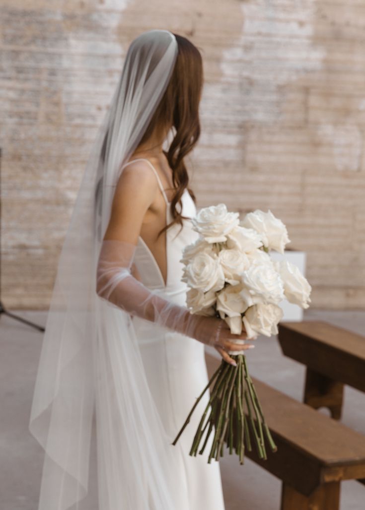 a woman in a white wedding dress holding a bouquet of flowers next to a bench