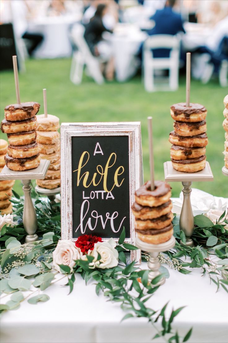 A wedding tables cape with greenery features a chalkboard sign with "A whole lotta love" written on it. Also on the table are several pedestals with multiple donuts on each pedestal, ready for guests to enjoy. Wedding Donut Dessert Table, Donut Dessert Table, Bridal Brunch Decorations, Donut Bar Wedding, Outdoor Wedding Backdrops, Donut Display, Wedding Donuts, Donut Dessert, Dessert Bar Wedding