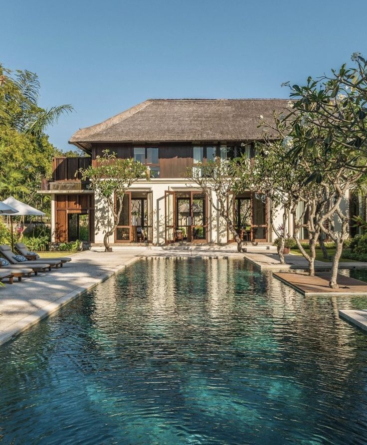 an outdoor swimming pool with lounge chairs and umbrellas in front of the house that is surrounded by trees