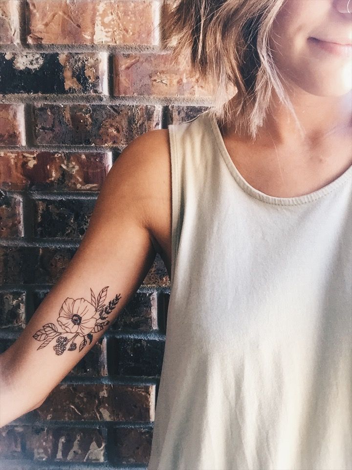 a woman with a flower tattoo on her arm next to a brick wall holding a cell phone