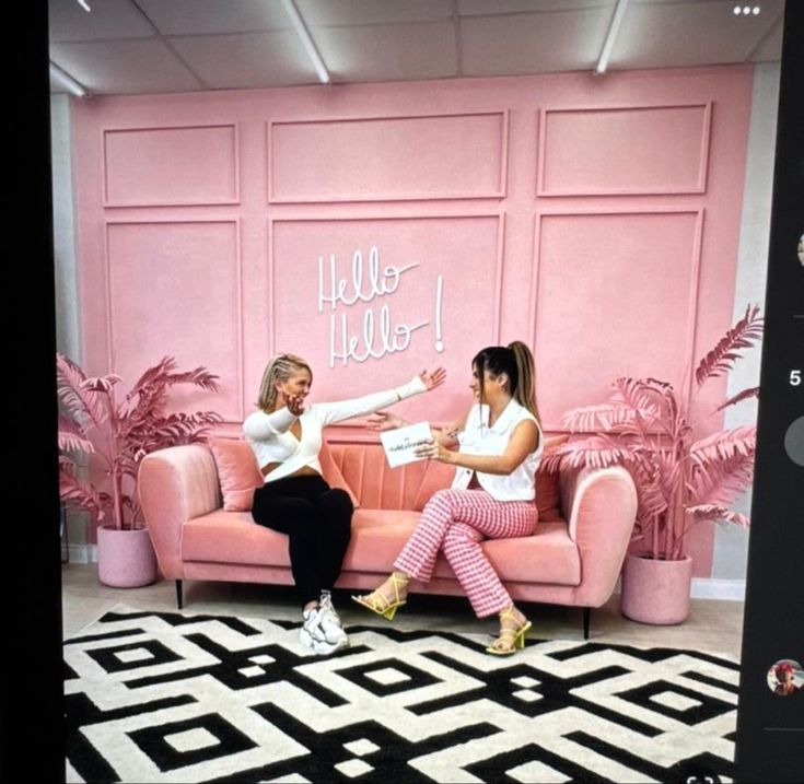 two women sitting on pink couches in front of a wall with hello hello written on it