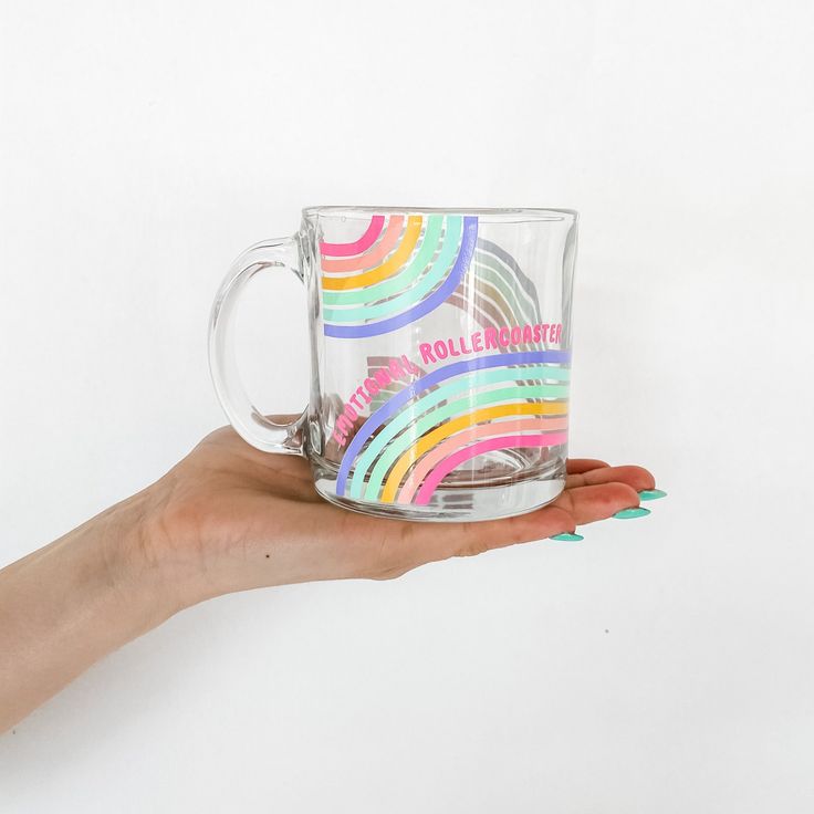 a hand holding a clear glass mug with rainbow designs on the bottom and inside it