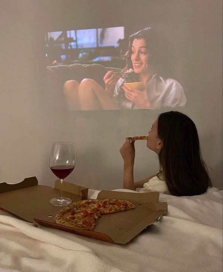 a woman sitting on a bed eating pizza and drinking wine in front of a tv