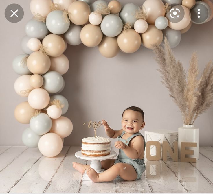 a baby sitting on the floor with a cake and balloons in front of him,