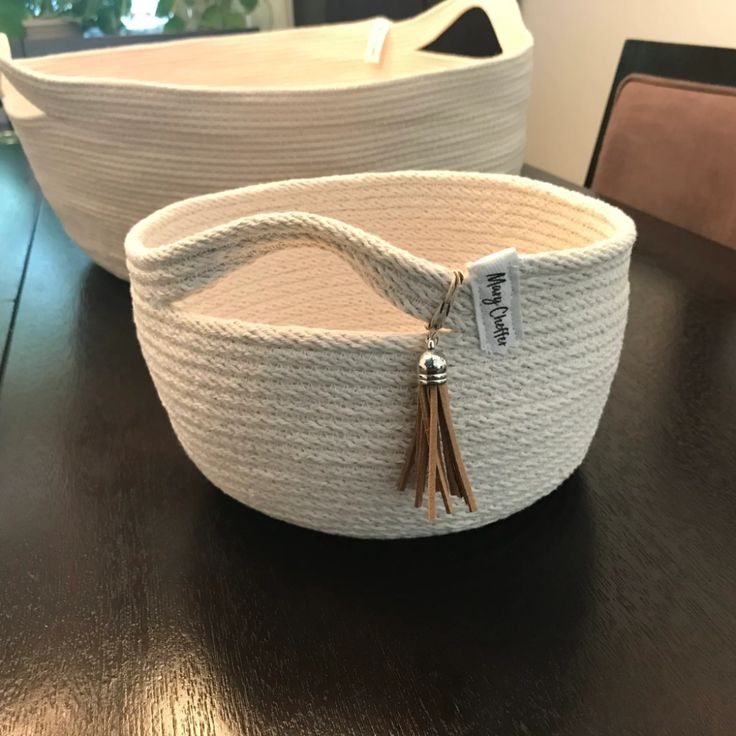 two white baskets sitting on top of a wooden table next to each other, one with a tassel hanging from it