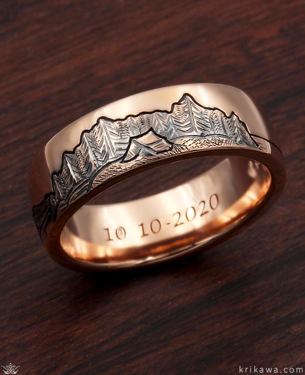 a gold wedding ring with mountains engraved on the front and sides, sitting on a wooden surface