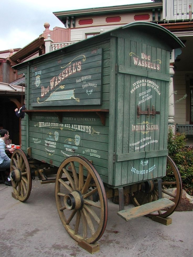 an old fashioned wooden wagon is parked in front of a building with people walking around it