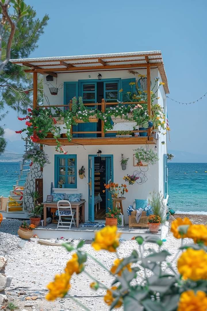 a white house with blue shutters and yellow flowers in front of the ocean on a sunny day