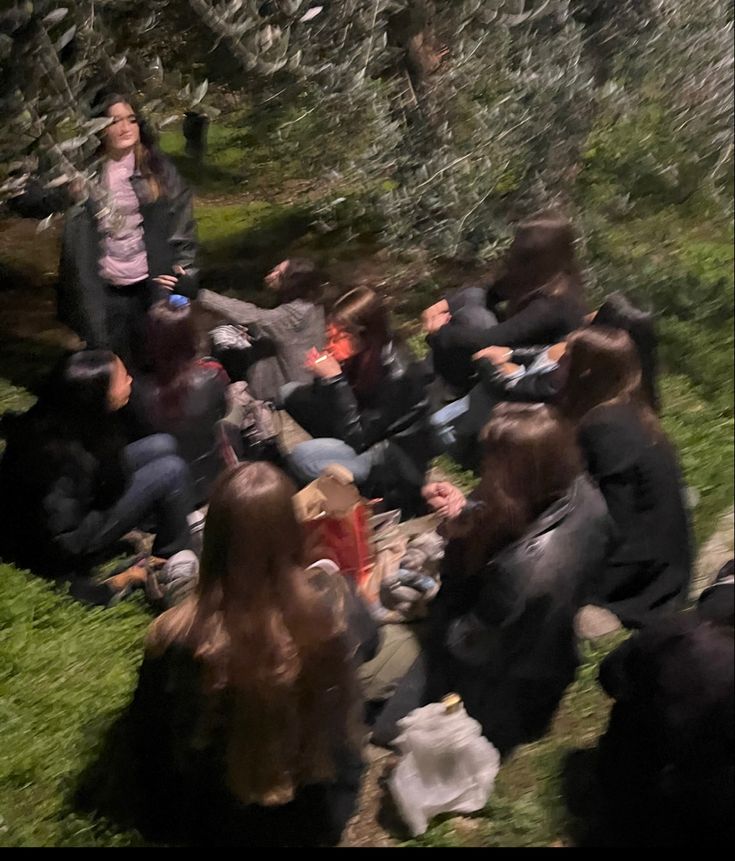 a group of people sitting on the ground in front of some trees and one person standing