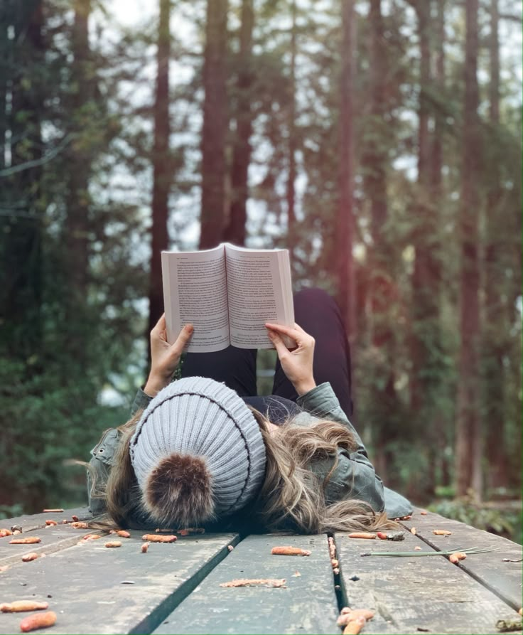 a woman laying on the ground reading a book in the woods with her head resting on an open book