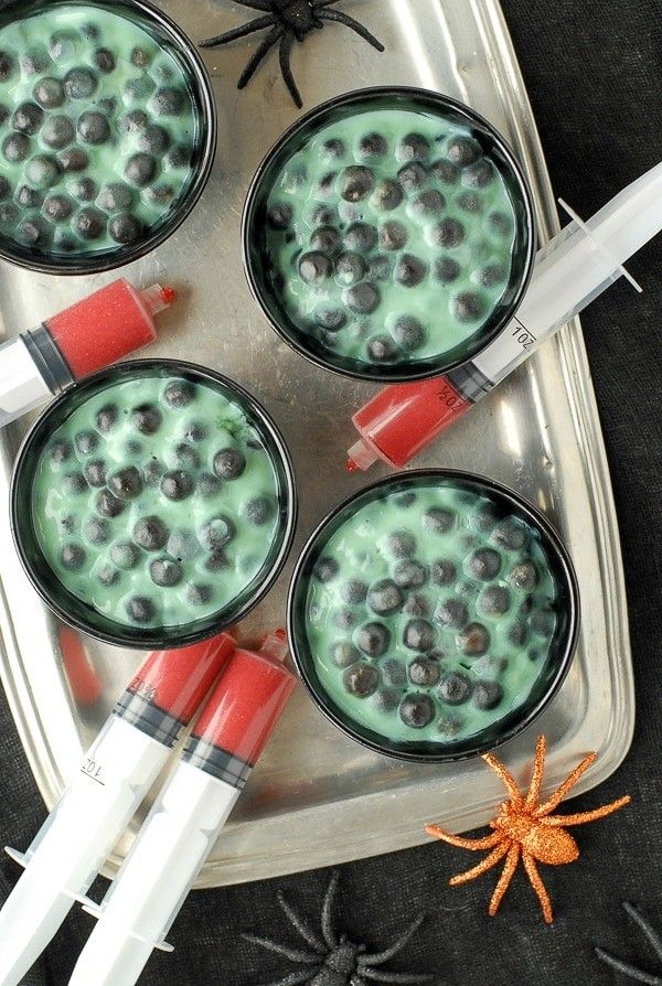 four glasses filled with green liquid and blueberries in front of a spider on the table