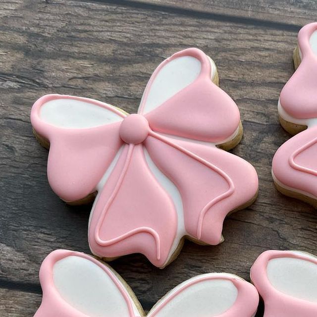 four decorated cookies with pink icing on a wooden surface, one is shaped like a bow