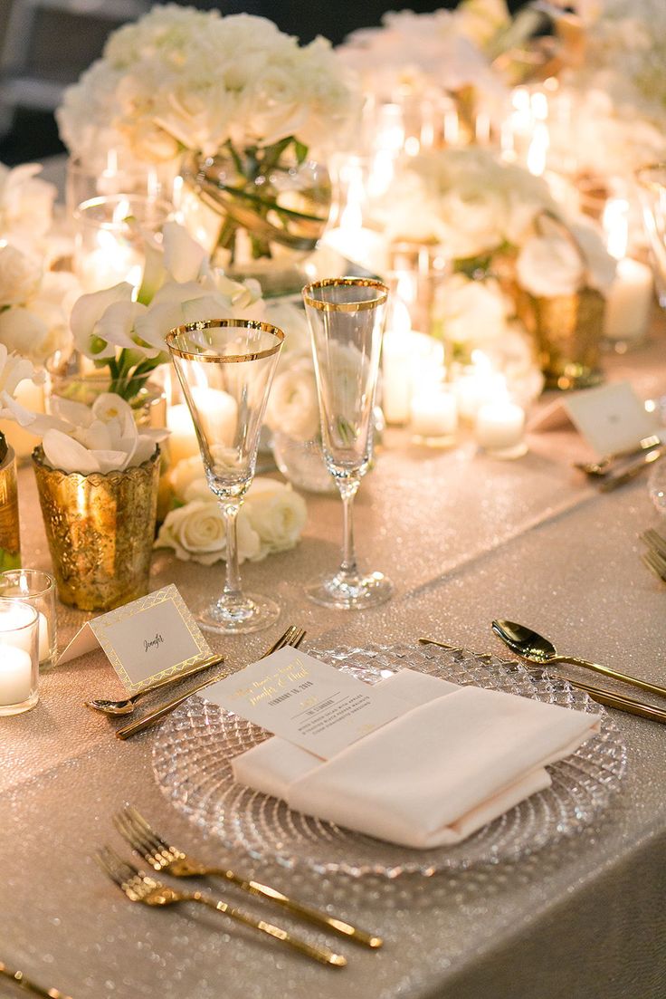 the table is set with white flowers and gold place settings, silver napkins and candles