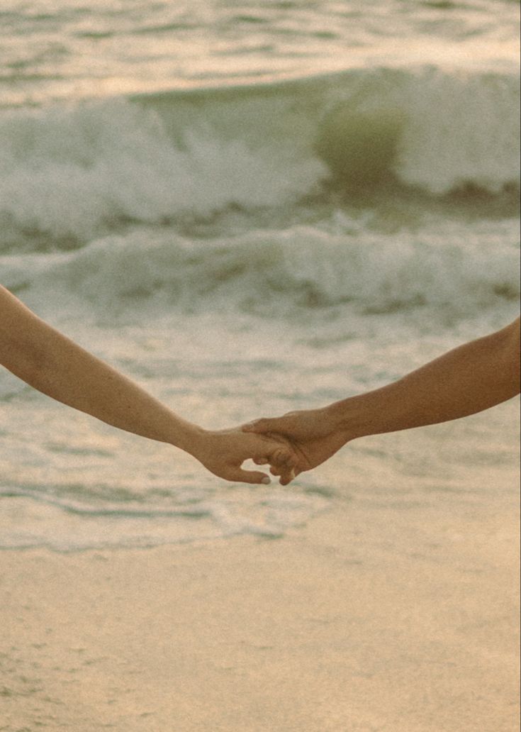 two people holding hands on the beach with waves crashing in the backgroound