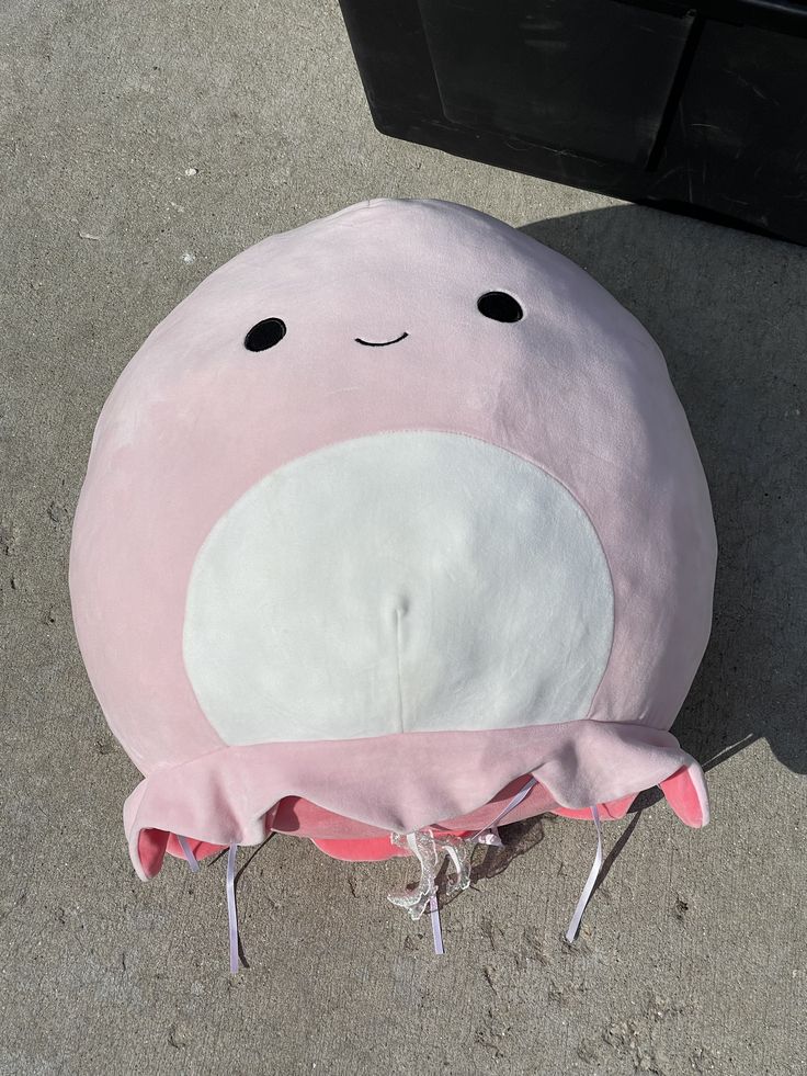 a pink and white pillow sitting on the ground next to a black trash can with its eyes closed