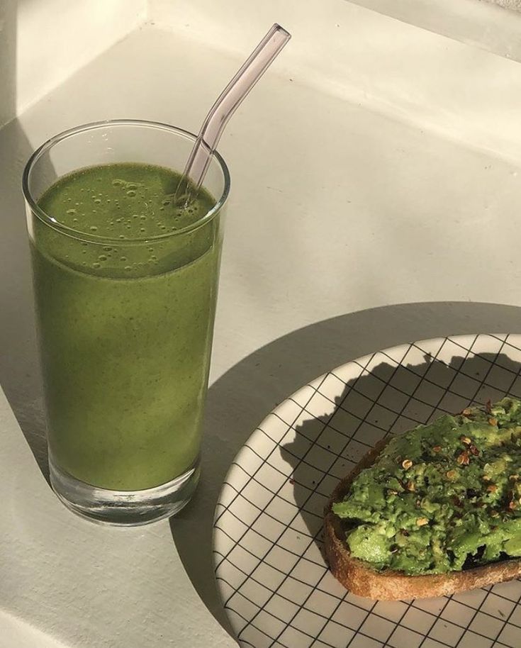 a glass of green smoothie next to a piece of bread on a white plate