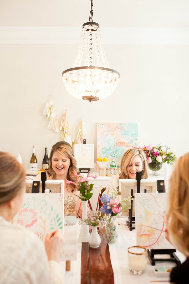 three women sitting at a table with flowers in vases and paintings on the wall