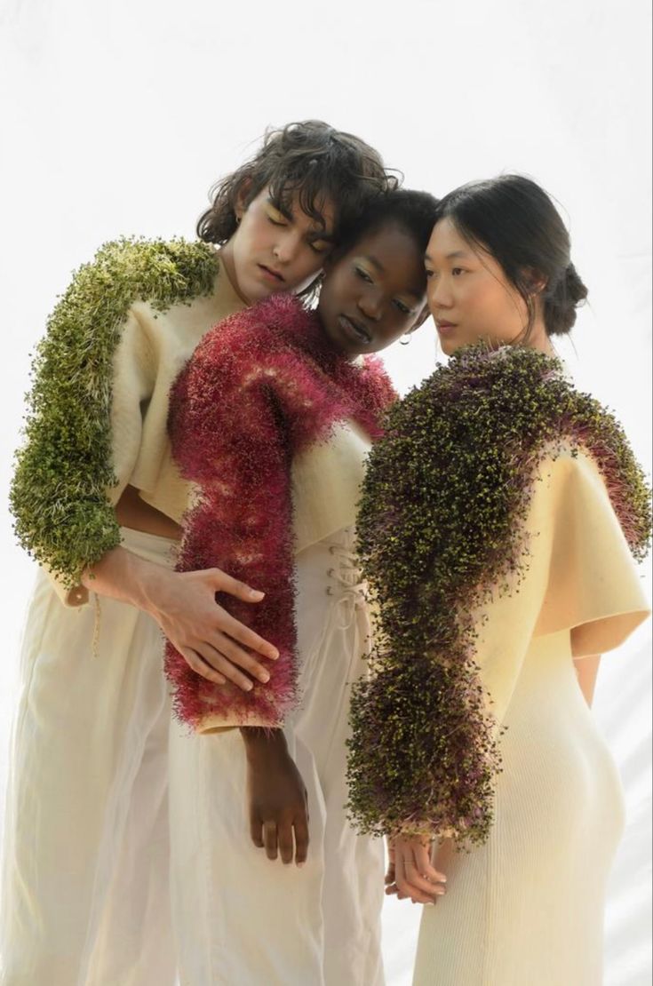 three women standing next to each other wearing white dresses and holding plants on their shoulders