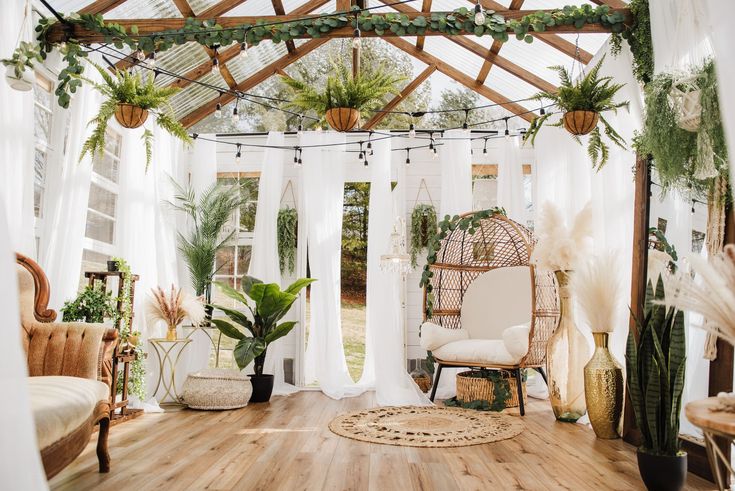 a room filled with lots of plants and hanging curtains on the ceiling, along with a wicker chair