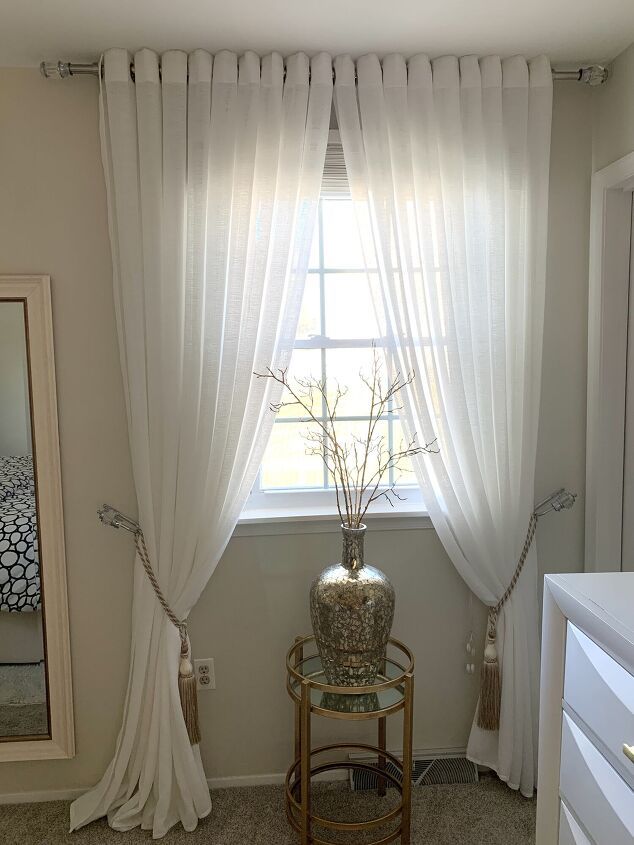 a white curtained window in a bedroom next to a table with a vase on it