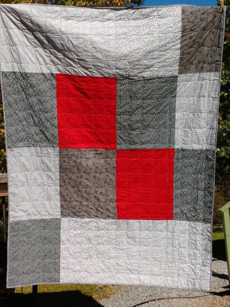 a red and grey quilt hanging from a clothes line in front of some trees on a sunny day