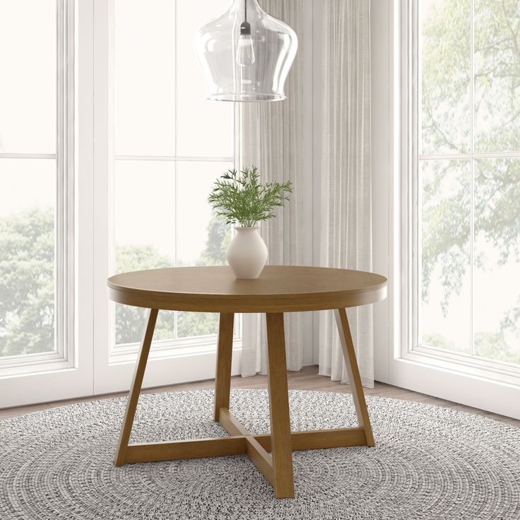 a round wooden table with a plant on it in front of two windows and a rug