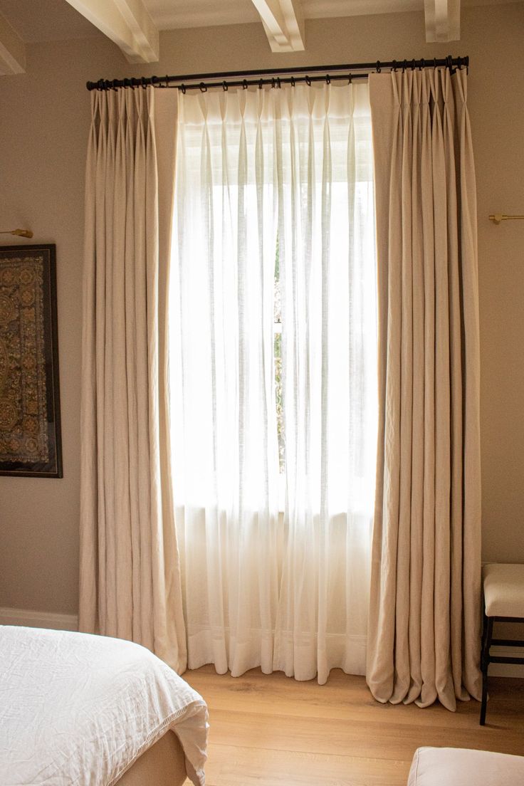 a bed sitting under a window next to a white curtain and chair in a bedroom