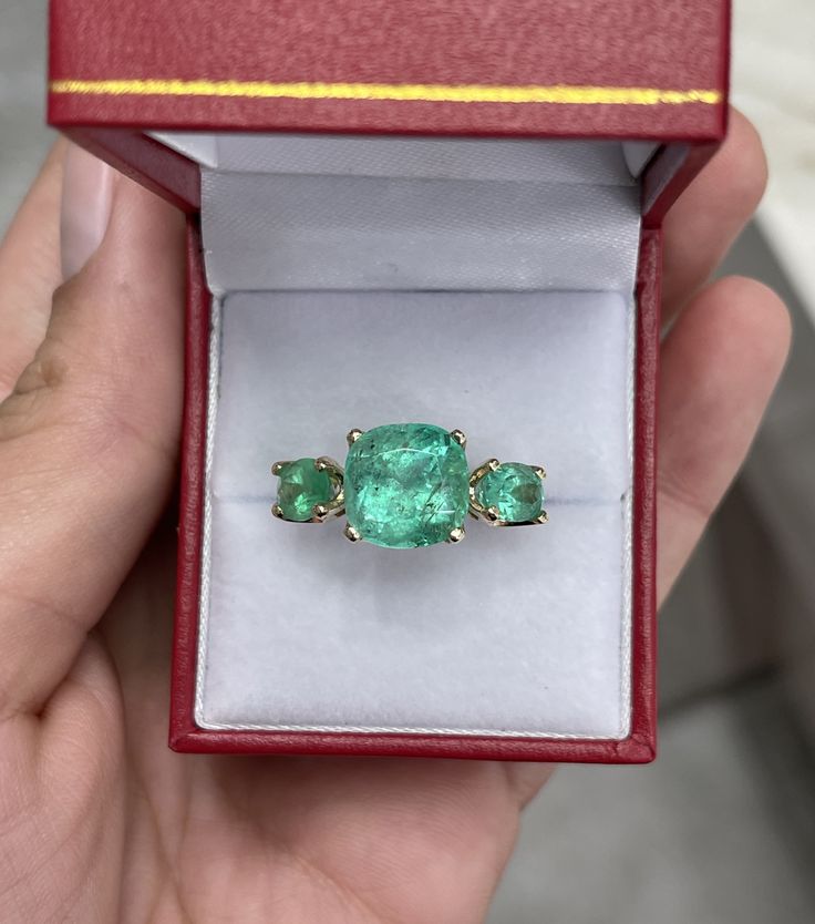 A Colombian emerald three stone ring. This bespoke ring design is crafted in gleaming 14K yellow gold, this ring features a 6.55-carat cushion cut in its center. Oval emeralds are flanked on the side creating an array of green. Simply gorgeous and one of a kind! Setting: Three-Stone Metal Purity: 14K Yellow Gold Center Stone: Emerald Shape: Cushion Cut Estimated Center Weight: 6.55-Carats Clarity: Semi-Transparent Color: Green Luster: Very Good Origin: Colombia Treatments: Natural Side Stones: E Luxury Three Stone Emerald Ring Gift, Luxury Three-stone Emerald Ring For Anniversary, Luxury Three Stone Emerald Ring For Anniversary, Luxury Gold Emerald Ring With Three Stones, Green Three Stone Birthstone Ring In 14k Gold, Luxury Three-stone Emerald Ring, Three Stone Emerald Promise Ring With Round Cut, Formal Three Stone Emerald Ring, Formal Emerald Three-stone Ring