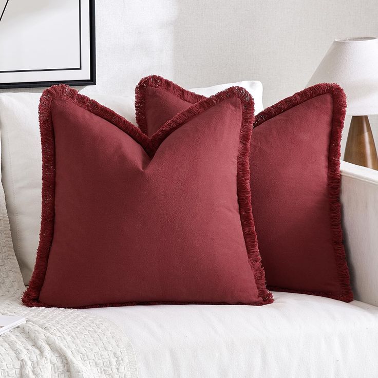 two red pillows sitting on top of a white couch next to a lamp and pictures