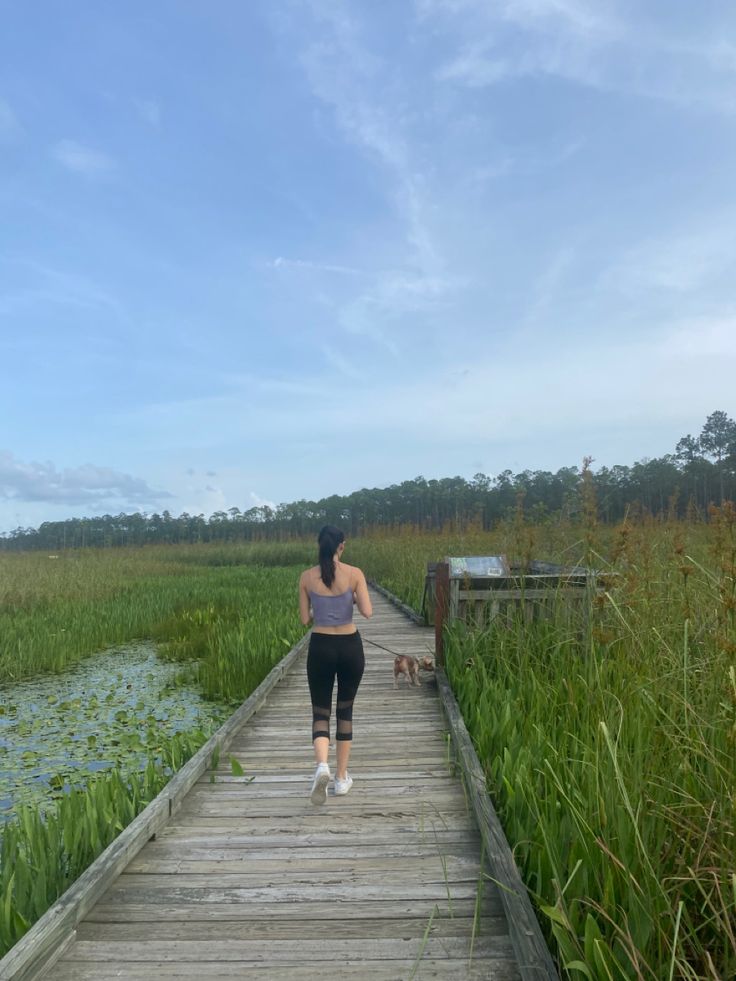 a woman walking across a wooden bridge next to a body of water and tall grass