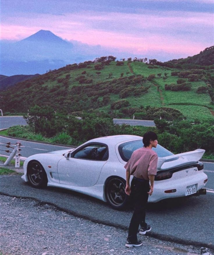 a man standing next to a white sports car on the side of a mountain road