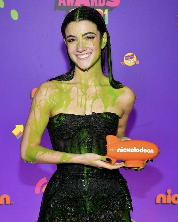 a woman with green paint on her face holding an orange blow dryer in front of a purple background