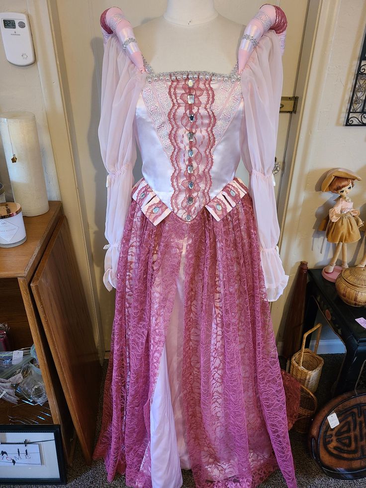 This gorgeous pink satin and mauve lace gown is perfect for that rennaisance faire, or for a stage play. It has just a touch of spatkle in the satin and is adorned with octagonal acrylic stones and induvidually knotted pearls. The ribbon has a pink embroidered floral look with metallic silver contrast. Thr sleeves have soft elastic and white satin bow detail. The back has larger grrommets in case you do stage plays and need to unlace quickly. The skirt hooks can be adjusted dependinb on the waist size. The small headpiece has matching lace and pearls for that added touch to complete the look.  Dress wilp fit a size 4 to 6 teen or small adult and is designed to be worn with or without a hoop skirt underneath.  This dress has 24 hrs of construction time put into it. Pink Lace Fitted Ball Gown, Elegant Pink Lace Ball Gown, Pink Elegant Ball Gown For Fancy Dress, Elegant Pink Ball Gown For Fancy Dress, Pink Elegant Ball Gown For Formal Event, Pink Fitted Gown For Costume Party, Fitted Pink Gown For Costume Party, Pink Satin Ball Gown For Debutante Ball, Fitted Pink Ball Gown With Lace Bodice