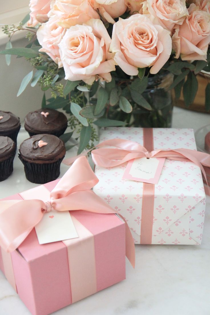 a vase filled with pink roses and cupcakes next to two wrapped gifts on a table