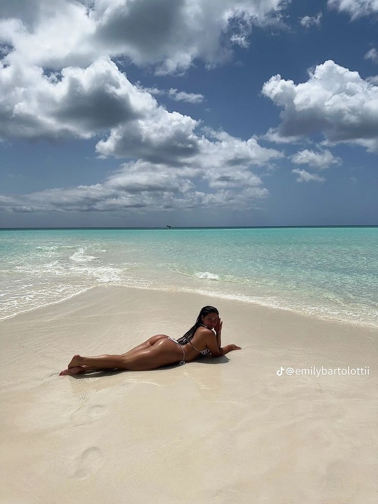 a woman laying on top of a sandy beach under a blue sky with white clouds