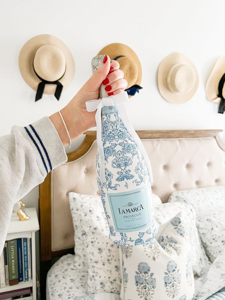 a woman holding a bottle of wine in front of a bed with hats on the headboard