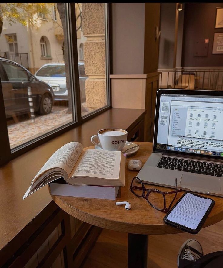 an open book sitting on top of a wooden table next to a laptop computer and cup of coffee