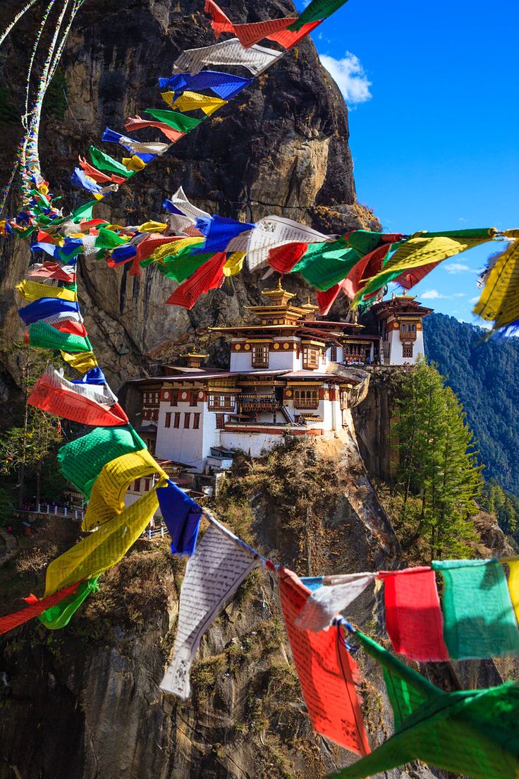 many colorful flags are flying in the air near a building on a mountain side with a cliff behind it